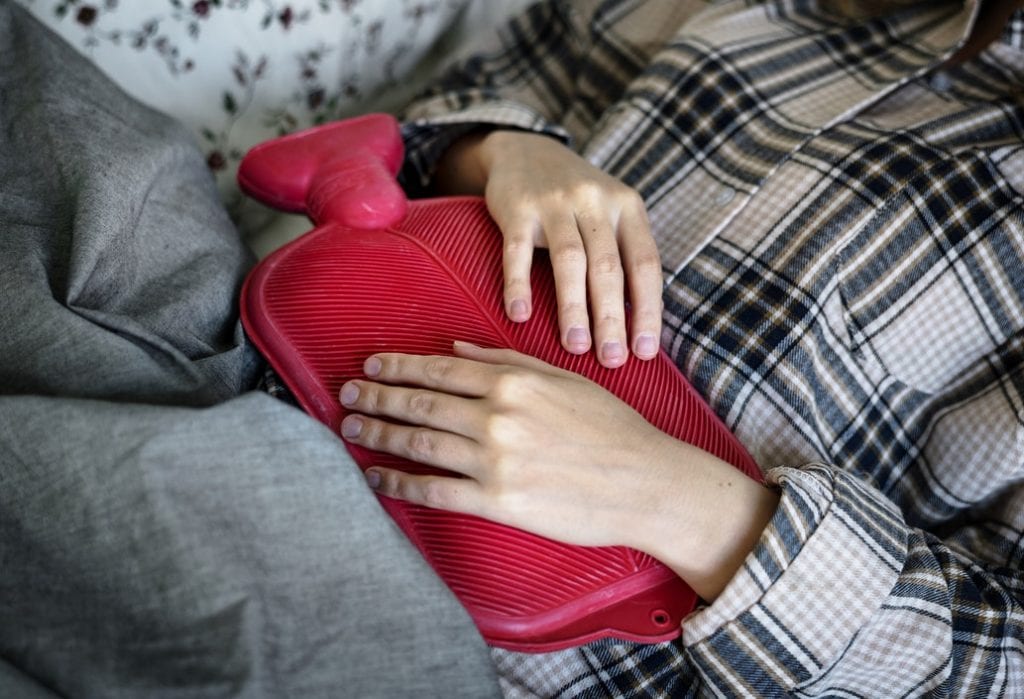 Woman holding a hot water bottle on her stomach having trouble with digestion. Digestion is a benefit of drinking more water. 