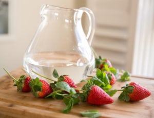 Big glass pitcher of water with strawberries laying around it on a work table in the kitchen. Strawberries are a great way to flavor water. 