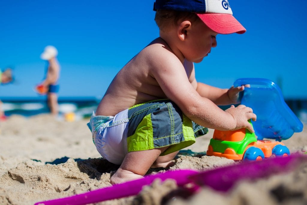2 year old boy with no shirt, trunks, and a baseball cap is playing on the beach with toys in the sand. 