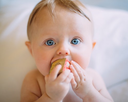 Baby with blue eyes chewing on block which shows you that toys for kids this age will end up in their mouth. 