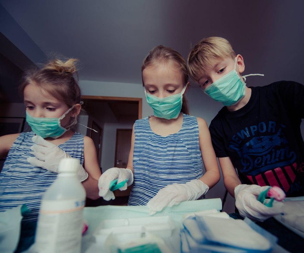 Three 4-5 year old kids with toys pretending to be doctor's and performing an operation. They all have on masks and gloves. 