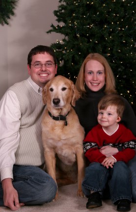 Husband wife son and a golden retriever taking a family holiday picture in front of the Christmas tree. 