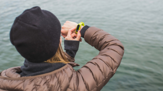 Woman in jacket standing by water looking at her yellow Garmin Vivofit fitness band.