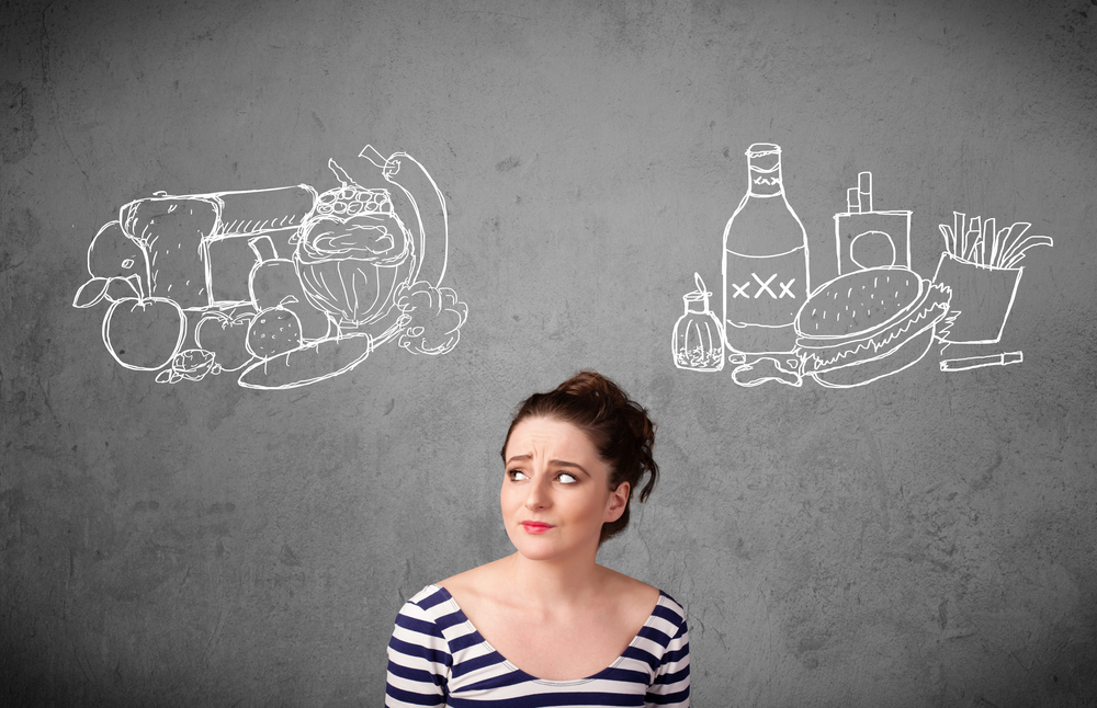 Woman thinking about Stress Eating and decinging between junk food or healthy food on displayed on the chalkboard behind her.
