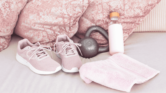 Workout shoes, kettle bell, water bottle, and workout towel sitting on a bed with pastel pink pillows.
